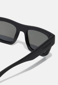Occhiali da sole neri con lenti scure, realizzati in plastica opaca, caratterizzati da un design geometrico contemporaneo e dal marchio inciso sull'asta.