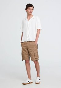 Camicia bianca a maniche corte con bottoni, pantaloni cargo color khaki con tasche laterali e sneakers bianchi con strisce nere.