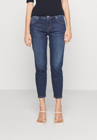 Tmavě modré skinny džíny s hladkým denimovým vzhledem, mají pět kapes, zapínání na knoflík a zip a ohrnutý lem. Džínsy jsou kombinovány s průhlednými sandály.