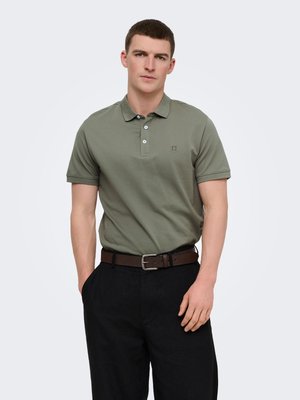 Jonge man draagt een olijfgroen poloshirt met drie knopen, zwarte broek, bruine riem, staat ontspannen tegen een witte achtergrond.
