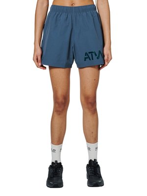 Person iført blå elastiske shorts med teksten "ATW", hvide sokker med mærket "HALO" og sorte sportssko, stående mod en hvid baggrund.