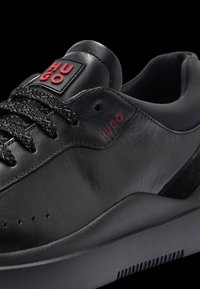 Sneakers en cuir noir avec une surface texturée, des accents de logo rouges et une semelle en caoutchouc foncé dotée d'ouvertures de ventilation.