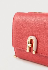 Κόκκινο πορτοφόλι από δερμάτινη υφή με χρυσή και εκρού μεταλλική λεπτομέρεια. Κλείνει με φερμουάρ στην κορυφή· συμπαγές ορθογώνιο σχήμα.