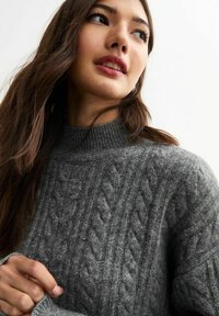 Maglione grigio a trecce con un collo alto. Presenta un motivo testurizzato e polsini a coste, evidenziando un design accogliente e lavorato a maglia.
