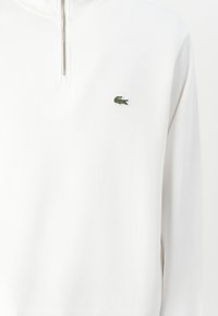 Vit pullover med kvartdragkedja, våffelstruktur och liten grön Lacoste-krokodil-logotyp på vänster bröst.