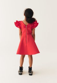 Robe rouge avec des manches à volants, texturée avec des logos. Présente une fermeture éclair au dos et une jupe évasée. Portée avec des bottines noires.