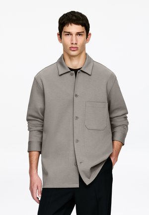 Młody mężczyzna ubrany w luźną szarą koszulę zapinaną na guziki z kieszenią na piersi, stojący z jedną ręką w kieszeni na jednolitym białym tle.