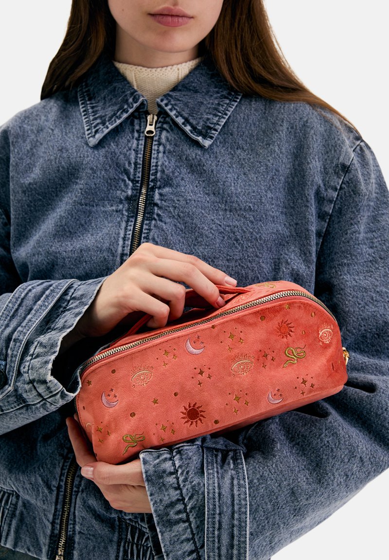 Kosmetyczka z zamszu w kolorze koralowym z zamkiem błyskawicznym, ozdobiona złotymi wzorami gwiazd, księżyców i słońc, trzymana w ręce.
