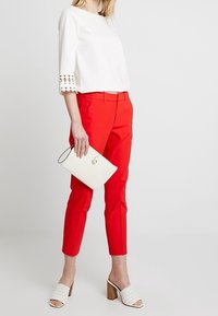 Haut blanc avec manches bordées de dentelle, pantalon rouge ajusté, pochette blanche et mules blanches tissées avec un talon bloc.