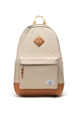 Béžový batoh s predným vreckom na zips, hnedým koženým spodkom a diamantovou nášivkou, a bielou štítkou s čiernym textom na pravom dolnom vrecku.
