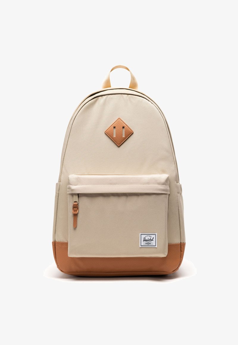 Sac à dos beige avec poche avant zippée, base en cuir marron et écusson en forme de diamant, ainsi qu'une étiquette blanche avec texte noir sur la poche inférieure droite.