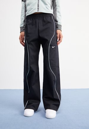 Person trägt schwarze Nike-Hose mit weißen Paspeln, elastischem Bund, Cargotasche, kombiniert mit weißen Sneakern und einer grauen Zip-Jacke.