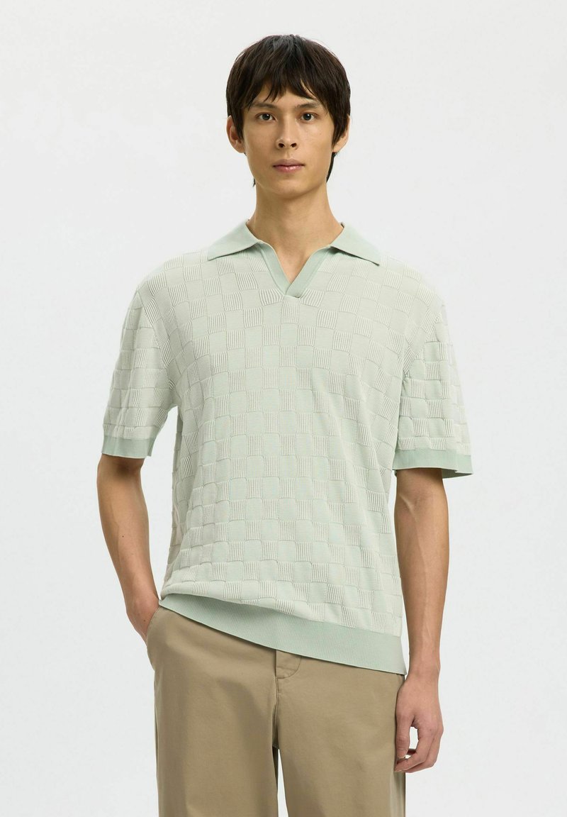 Jeune homme debout, la main dans la poche, portant un polo vert clair texturé à manches courtes et un pantalon beige sur un fond uni.