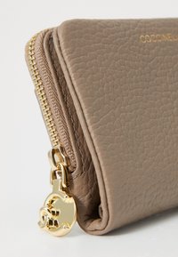 Brown leather wallet featuring a textured finish, gold zipper hardware, and a round charm pendant attached to the zipper pull.