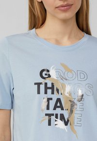Lichtblauwe katoenen T-shirt met een grafisch ontwerp met de tekst "GOEDE DINGEN HEBBEN TIJD NODIG," aangevuld met zwarte, gouden en zilveren tinten.