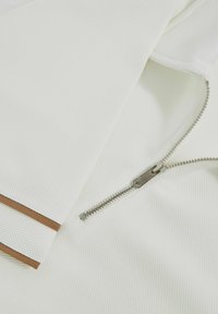 Biely textil s textúrovaným povrchom, s kovovým zipsom v leštenej striebornej farbe a hnedým akcentom na okraji.