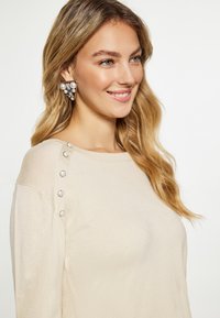Beige Langarmoberteil mit dekorativen Knöpfen an der Schulter und glatter Textur. Große Perlenohrringe vervollständigen den Look.