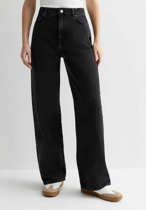 Persoon met een zwarte, losvallende jeans met hoge taille, gecombineerd met witte sneakers met beige accenten, staand tegen een effen lichte achtergrond.