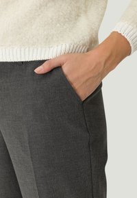 Szare dopasowane spodnie o gładkiej fakturze, z bocznymi kieszeniami. Ręka modelki opiera się w lewej kieszeni, a na sobie ma kremowy sweter z dzianiny.