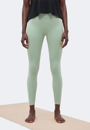 Osoba stoi boso na beżowej macie do jogi, ubrana w jasnooliwkowe legginsy i czarny krótki top na jednolitym jasnym tle.