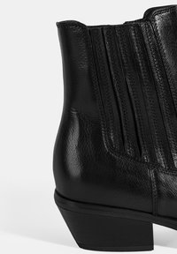 Botte de cheville en cuir noir avec une finition texturée, plis verticaux décoratifs et un petit talon empilé. La vue de profil latéral met en valeur les détails du design.