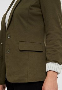 Olivgrön blazer i texturerat tyg, med två knappar, en enkel ficka och randiga manschetter som kikar fram under ärmarna.