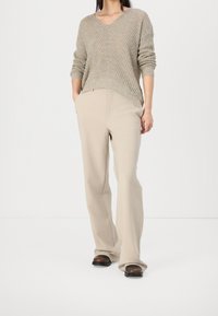 Pull en tricot beige avec un col en V et une texture côtelée, associé à un pantalon large crème et des sabots marron, sur fond blanc.