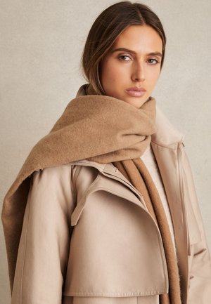 Ženska, oblečena v bež usnjeno jakno in velik svetlo rjav volnen šal, gleda v kamero na enostavni nevtralni ozadju.