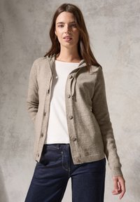 Beige stickad cardigan med huva, framknappar, ribbade ärmslut och strukturerade ärmar, buren över en vit topp och blå jeans.