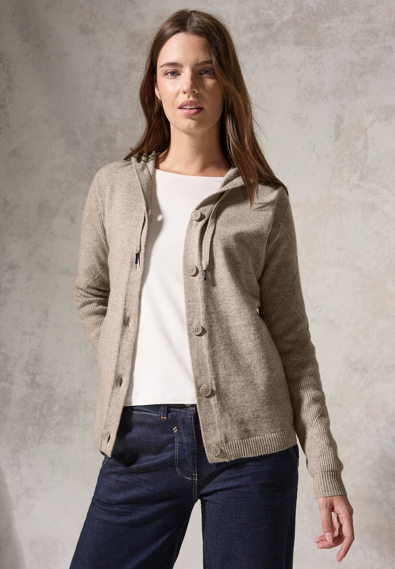 Beige stickad cardigan med huva, framknappar, ribbade ärmslut och strukturerade ärmar, buren över en vit topp och blå jeans.