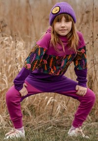 Fioletowy i różowy dres z bluzą w liściasty wzór i jednolitymi spodniami, w połączeniu z fioletową czapką z naszywką z uśmiechniętą buźką.