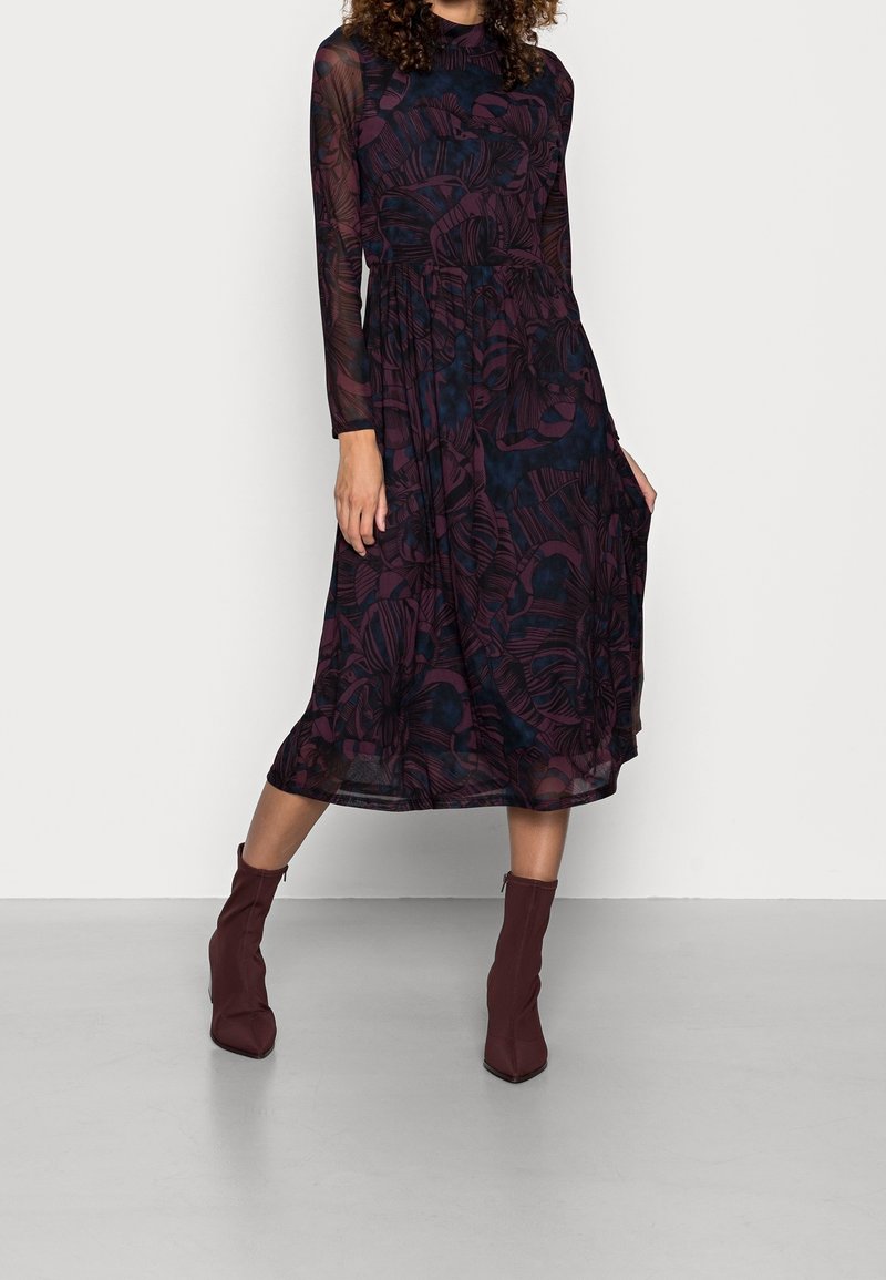 Femme portant une robe midi fleurie bleu marine et bordeaux transparente à manches longues avec des bottines bordeaux, debout sur un sol gris.