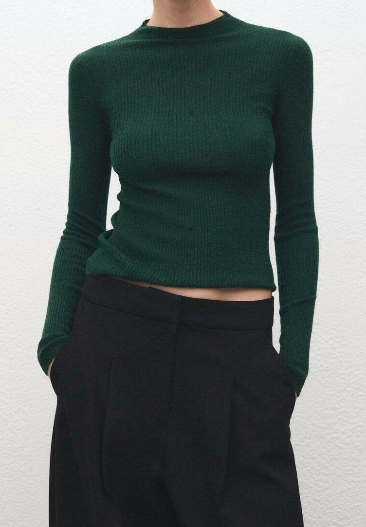Personne portant un haut à manches longues côtelé vert foncé et un pantalon noir taille haute à jambes larges, mains dans les poches, sur un fond clair uni.