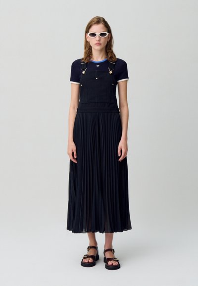 Salopette en denim noir avec des bretelles ajustables sur un haut ajusté en mailles bleu marine, associée à une jupe plissée noire et des sandales noires avec des accents dorés.