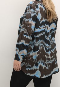 Brun og blå oversized skjorte med et bølget mønster, lange ærmer og en struktureret stof; buet kant og knapdetaljer ved ærmerne.