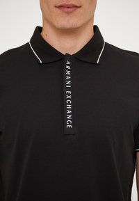 Polo noir avec un col blanc contrastant et une étiquette verticale portant l'inscription "ARMANI EXCHANGE". Texture de tissu lisse.