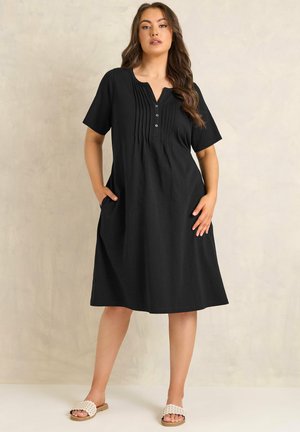 Kvinde iført en knælængde sort kjole med pintuck-detaljer og knapper, stående med hænderne i lommerne og hvide slip-on sandaler.