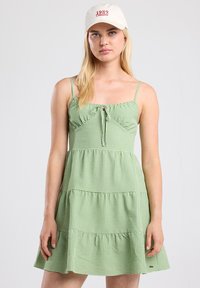 Vihreä ruudullinen mekko, jossa on istuva yläosa, etuosan solmitys, kerroksellinen rypytyshelma ja ohuet hihnat. Yhdistetty beigen väriseen lippikseen, jossa on punainen teksti.