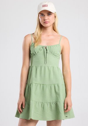 Robe vichy verte avec un corps ajusté, détail à nouer à l'avant, ourlet à volants en niveaux et fines bretelles. Portée avec une casquette beige affichant un texte rouge.