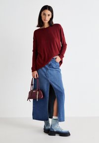 Bordeaux gebreide trui, blauwe denim maxirok met een zijsplit, lichtblauwe veterschoenen en een bordeaux handtas met kettingdetail.