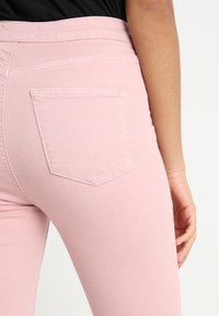 Vaqueros de mezclilla rosa con un diseño ajustado, que cuentan con un bolsillo trasero y costuras visibles a lo largo de la cinturilla. La textura parece suave y elástica.
