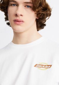 Camiseta de algodón blanca con un pequeño logo en naranja y amarillo de "Dickies" en el lado izquierdo del pecho. Cuello redondo y textura suave con un corte relajado.