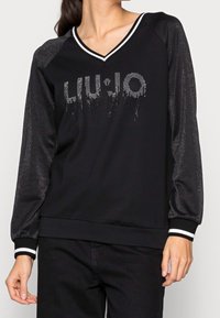 Kobieta w czarnej bluzie z dekoltem w serek i błyszczącymi rękawami oraz napisem "LIU•JO" z kryształków na piersi, w zestawie z czarnymi spodniami.
