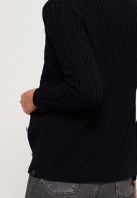 Navy-blauer Strickpullover mit gerippter Textur, langen Ärmeln und einem schmalen Bund, mit einem kleinen Markenlabel an der Seite.