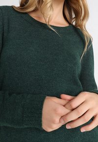 Mörkgrön stickad tröja med rundad halsringning, ribbade manschetter och en mjuk textur, med en slimmad passform. Händerna är knäppta fram.