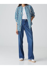 Koszula zapinana na guziki w niebiesko-beżowy wzór falisty, zestawiona z spodniami dżinsowymi o wysokim stanie w kolorze niebieskim. Model nosi białe buty z niebieskimi paskami.