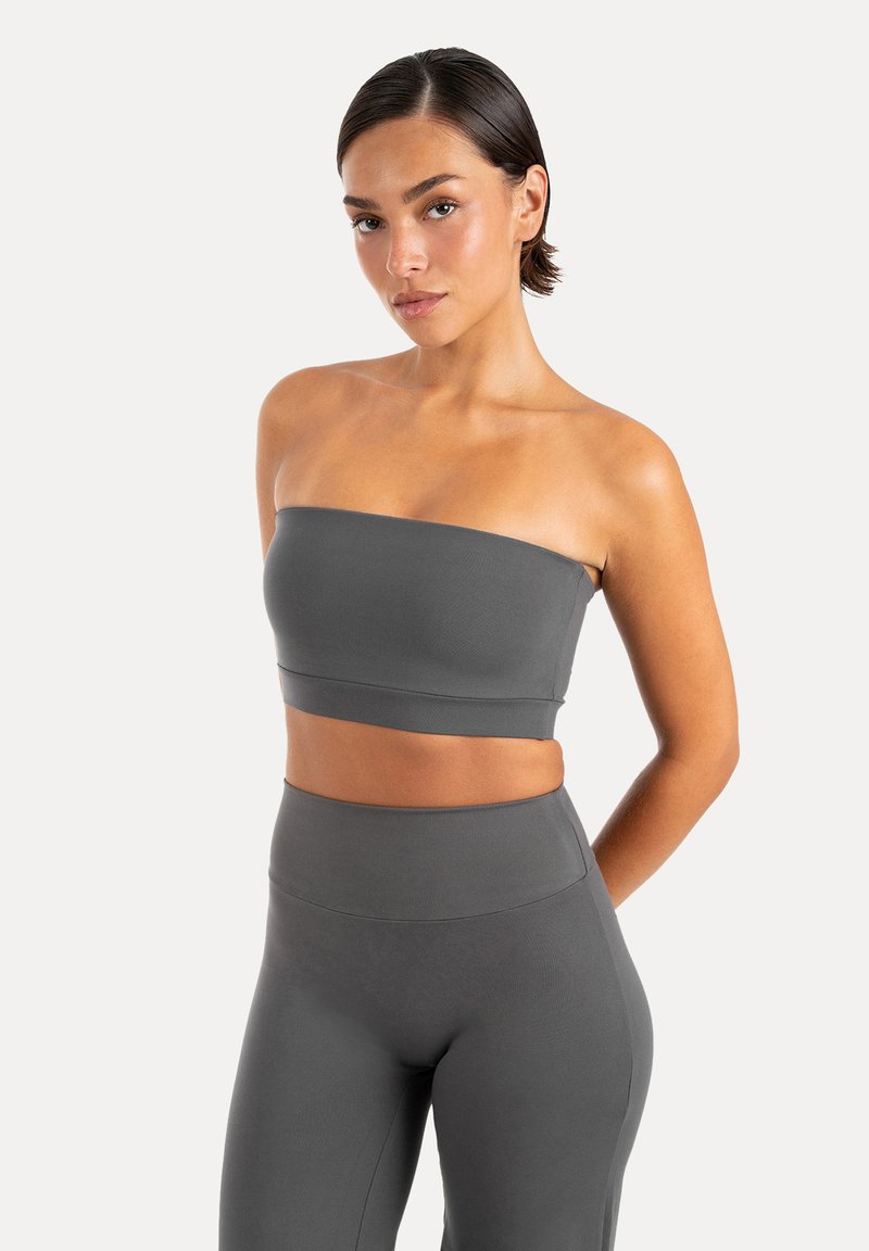 Femme aux cheveux courts et foncés portant un top court gris sans bretelles et un legging taille haute, debout devant un fond blanc uni.