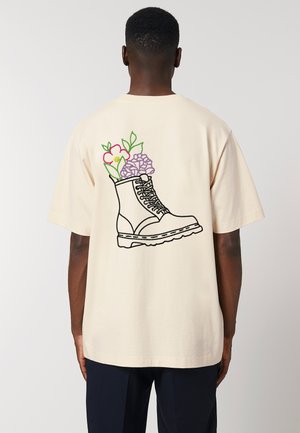 T-shirt couleur crème avec un graphique d'une botte contour noir remplie de fleurs colorées au dos. Manches courtes, coupe décontractée.