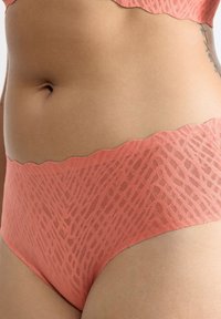 Close-up van een torso met koraalroze kanten ondergoed met zigzagpatroon en gegolfde randen, waarbij een kleine tatoeage aan de zijkant zichtbaar is.
