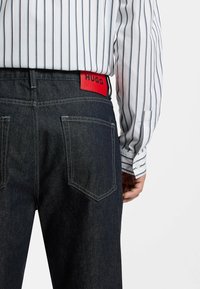 Persona che indossa jeans di denim scuro con un'etichetta rossa HUGO sulla vita e una camicia bianca con strisce verticali nere.